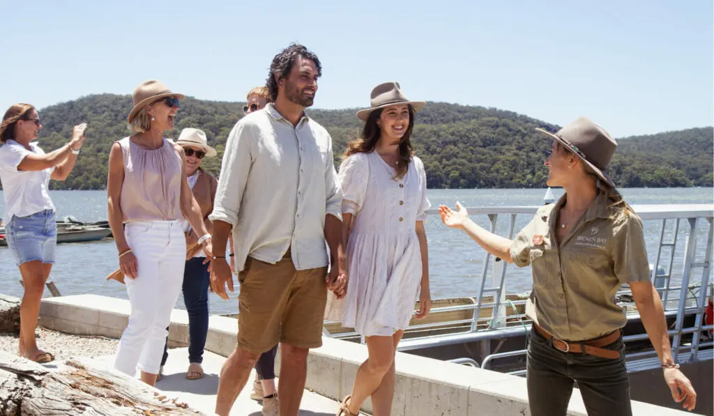 A tour guides takes her guests on a Farm Cruise Tour, leading them to the tour boat at the Shellar Door. Behind them is the Hawkesbury River and the landmasses with beautiful greenery on them.